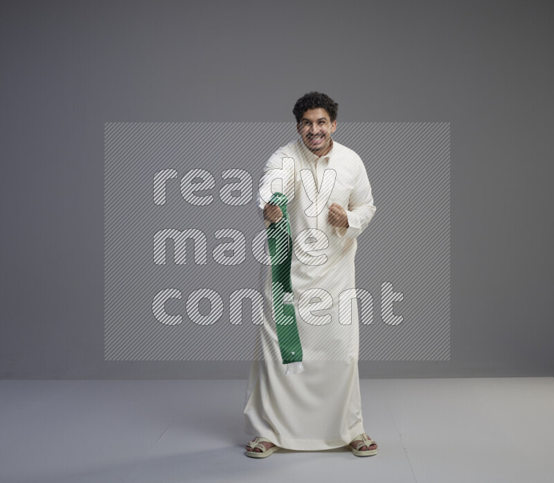 A Saudi man standing wearing thob holding Saudi flag scarf on gray background