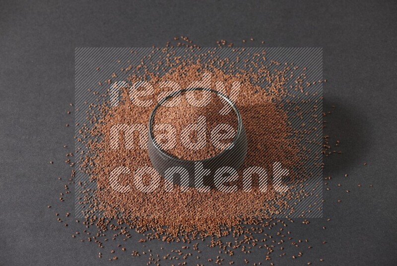 A black pottery bowl full of garden cress seeds surrounded by seeds on a black flooring