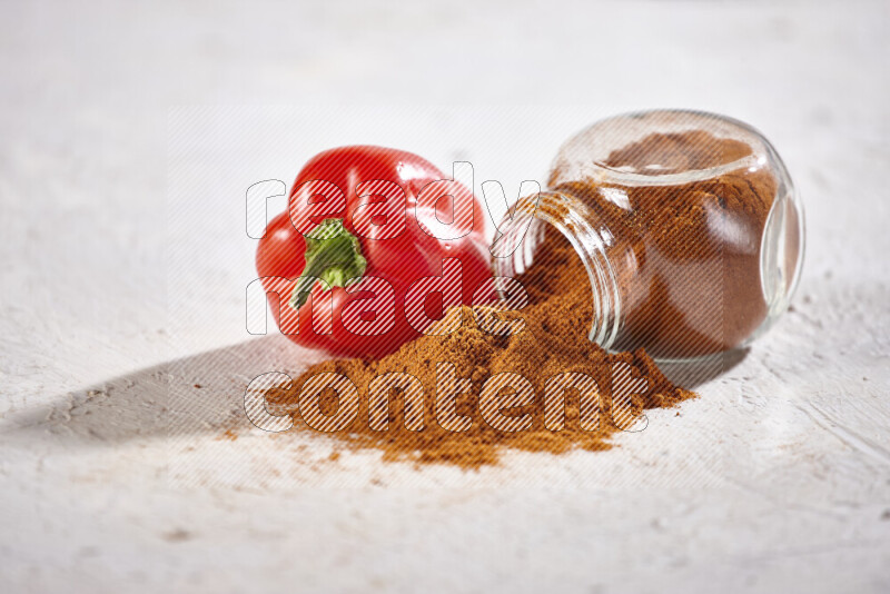 A glass jar full of ground paprika powder flipped with some spilling powder on white background
