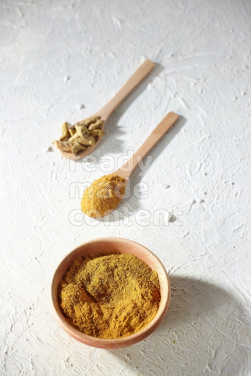 A wooden bowl full of turmeric powder and 2 wooden spoons full of dried turmeric whole finger and turmeric powder on textured white flooring