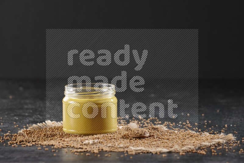 A glass jar full of mustard paste set on a burlap piece with a wooden spoon full of mustard seeds on a textured black flooring