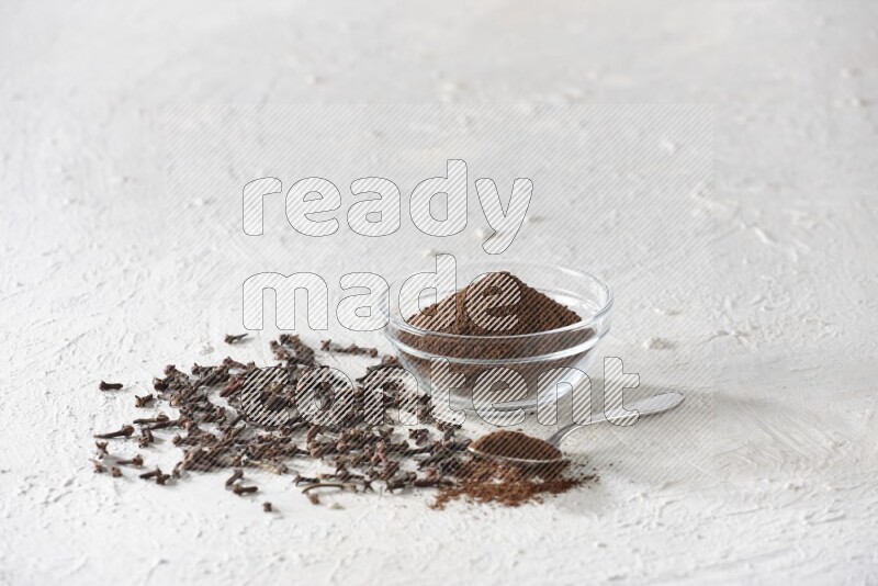 A glass bowl and a metal spoon full of cloves powder with cloves grains spread on a textured white flooring