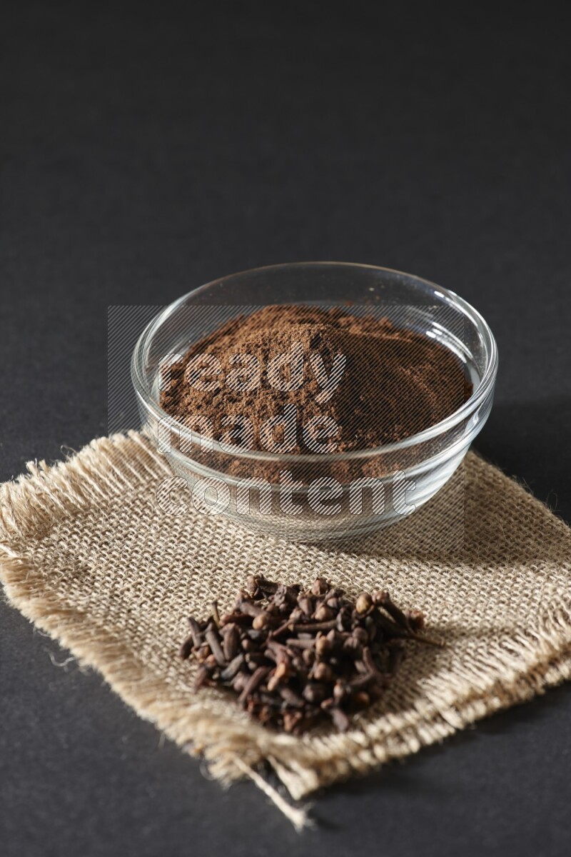 A glass bowl full of cloves powder with cloves grains on a burlap piece on a black flooring