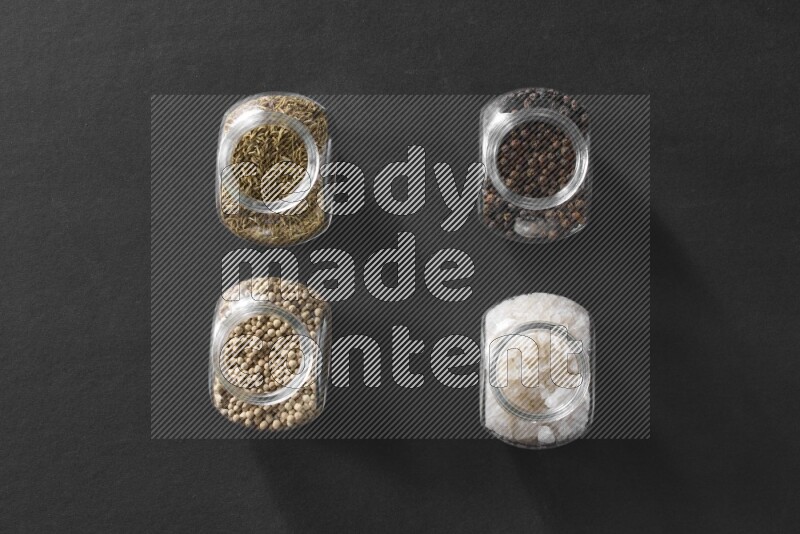 4 glass spice jars full of salt, black peppers, white peppers and cumin on black flooring