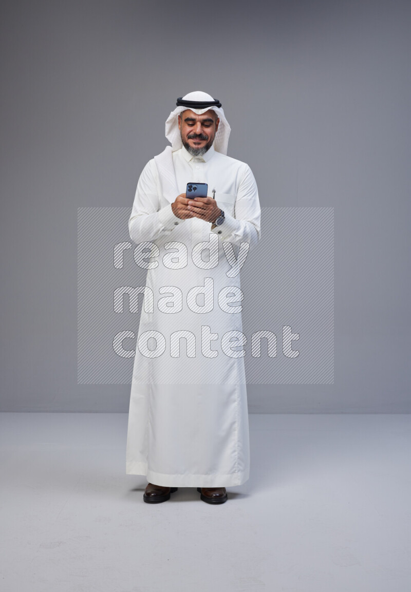 Saudi man Wearing Thob and white Shomag standing texting on phone on Gray background