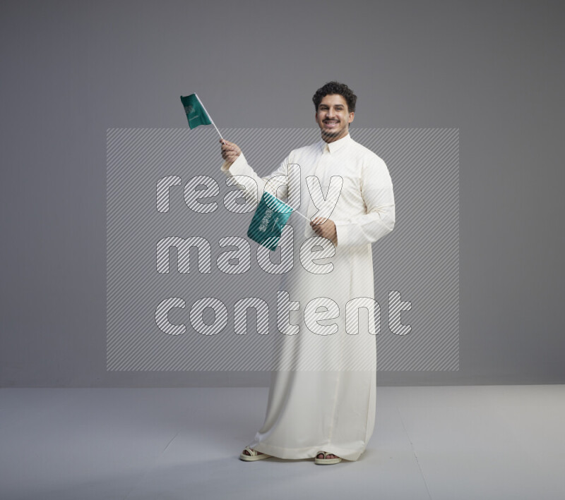 A Saudi man standing wearing thob holding small Saudi flag on gray background