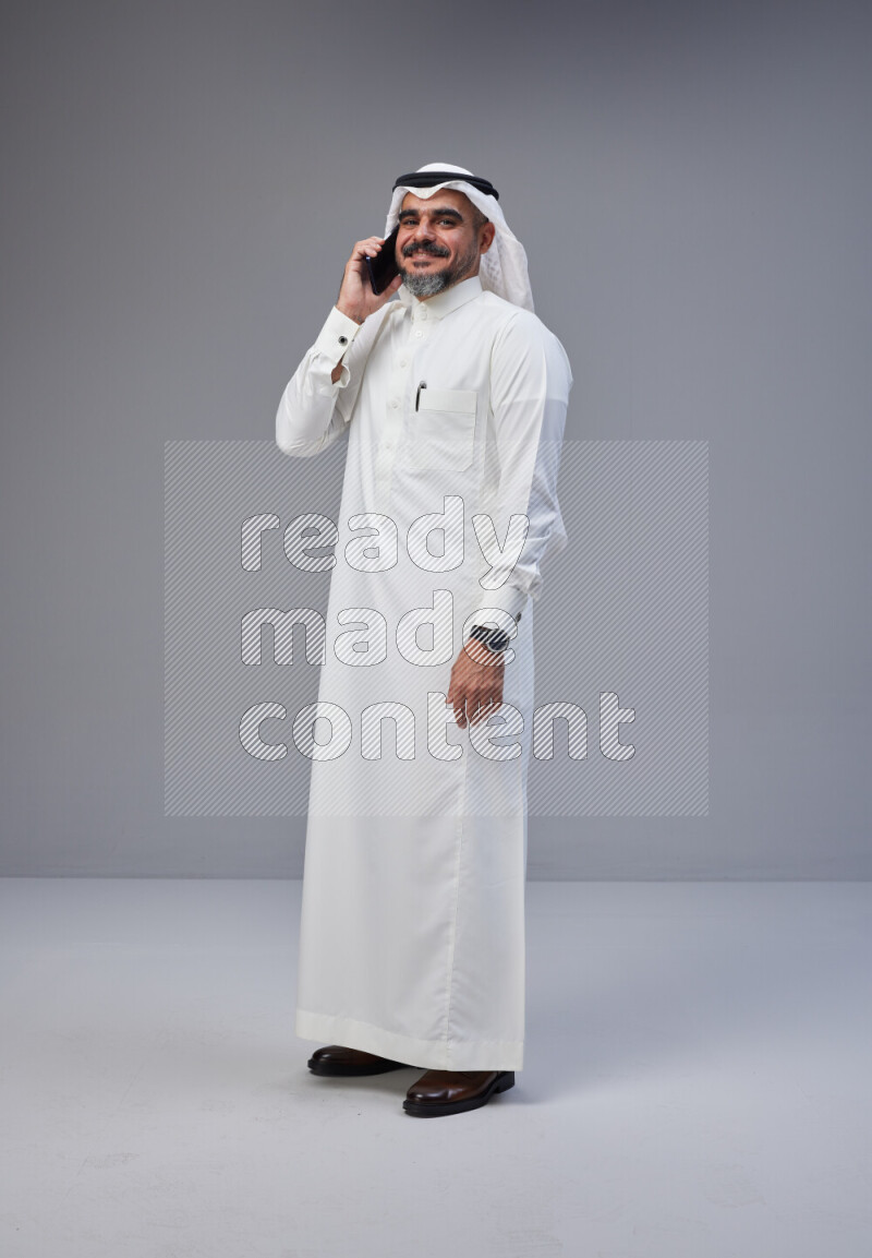Saudi man Wearing Thob and white Shomag standing talking on phone on Gray background