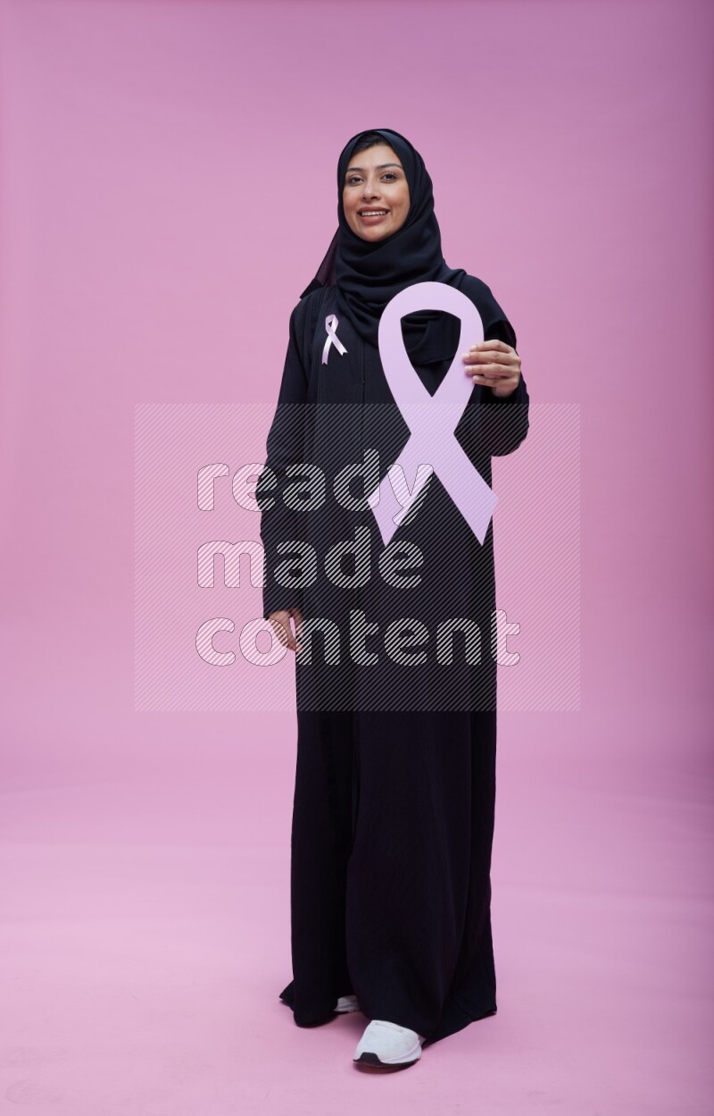 Saudi woman wearing pink ribbon on abaya standing holding awareness ribbon on plain pink background
