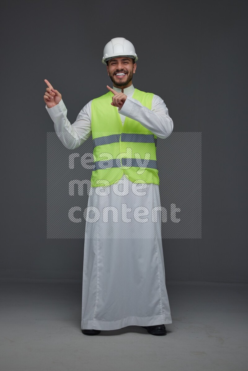 A Saudi man wearing Thobe with a yellow safety vest and white helmet standing and pointing different angles eye level on a gray background