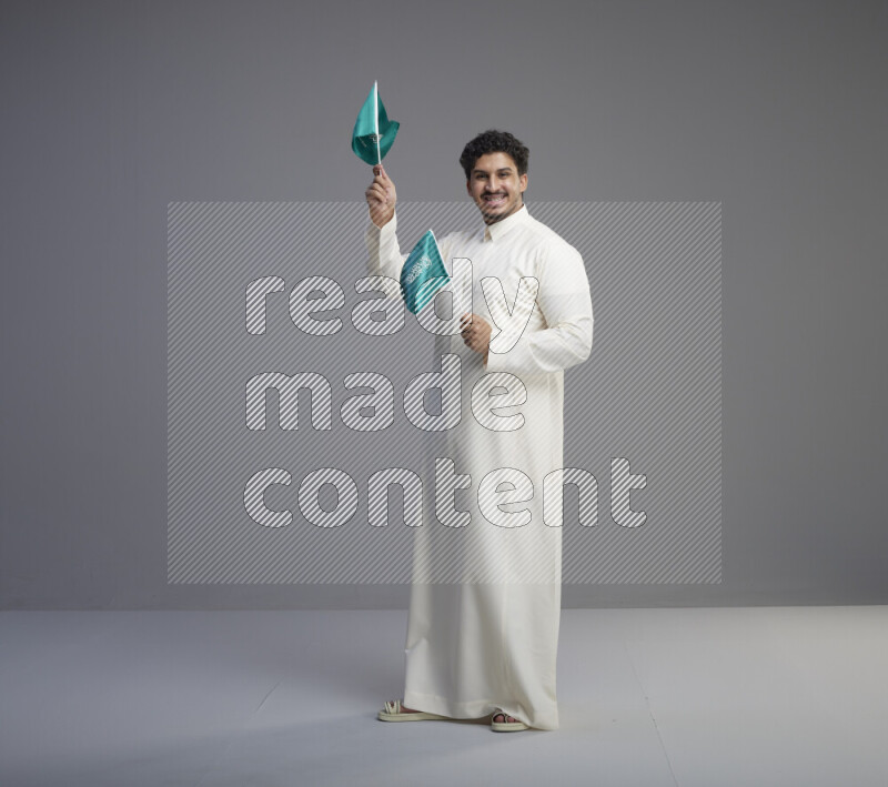 A Saudi man standing wearing thob holding small Saudi flag on gray background