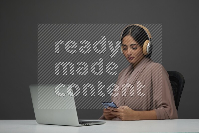 A Saudi woman Sitting on her desk Typing on her laptop wearing a headset  on a Gray Background wearing Brown Abaya