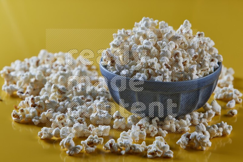 A blue pottery bowl full of popcorn with popcorn beside it on a yellow background in different angles