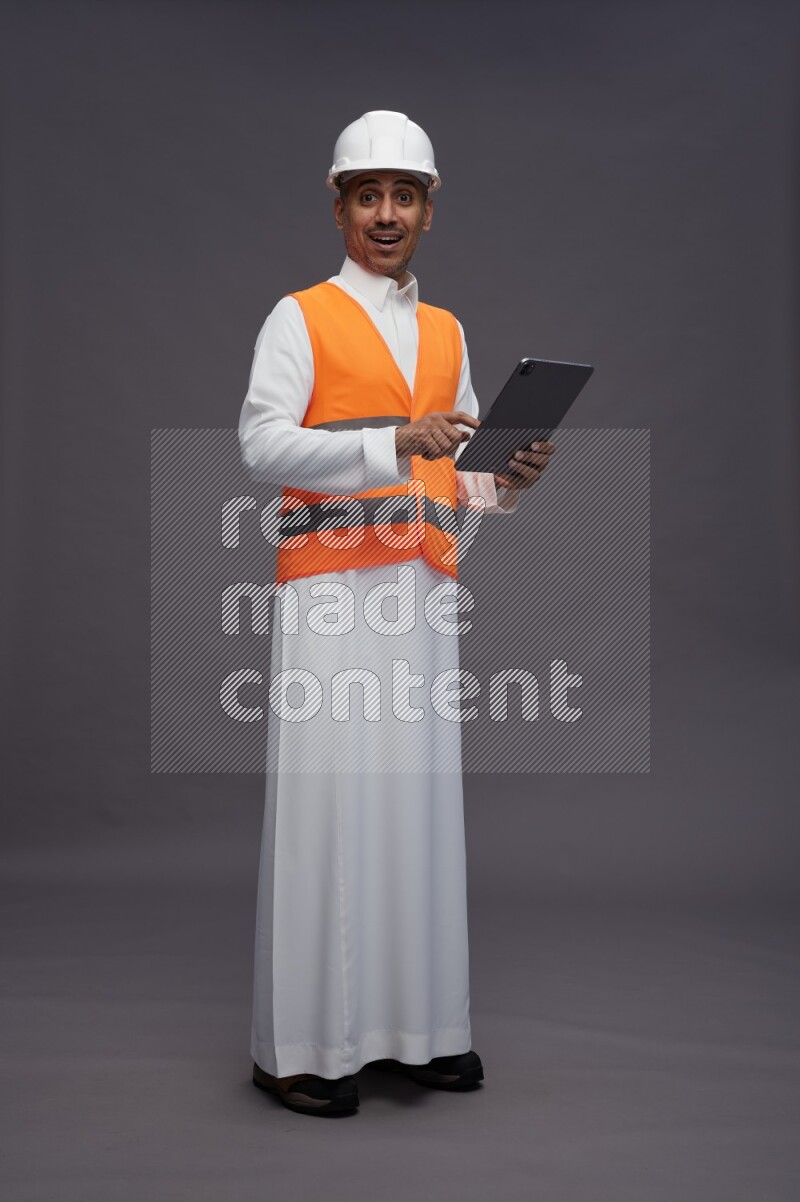 Saudi man wearing thob with engineer vest standing working on tablet on gray background
