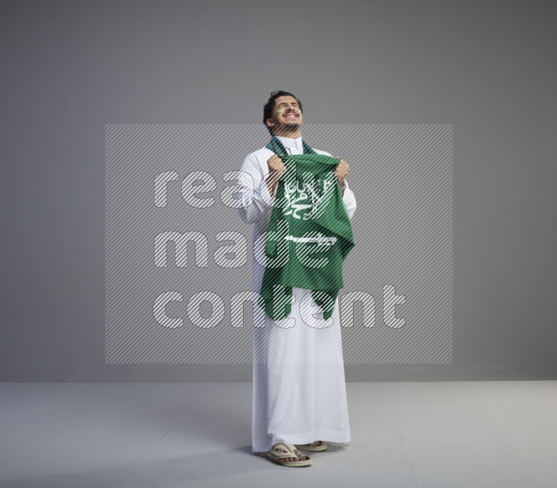 A Saudi man standing wearing thob with face painting wrapping Saudi flag scarf and holding big Saudi flag on gray background