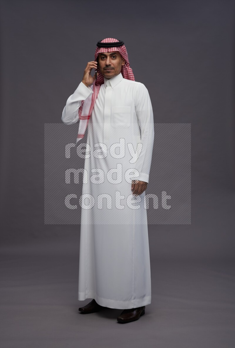 Saudi man wearing thob and shomag standing talking on phone on gray background
