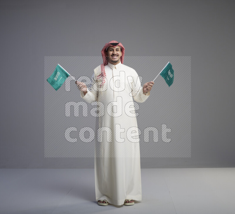 A Saudi man standing wearing thob and red shomag raising small Saudi flag on gray background