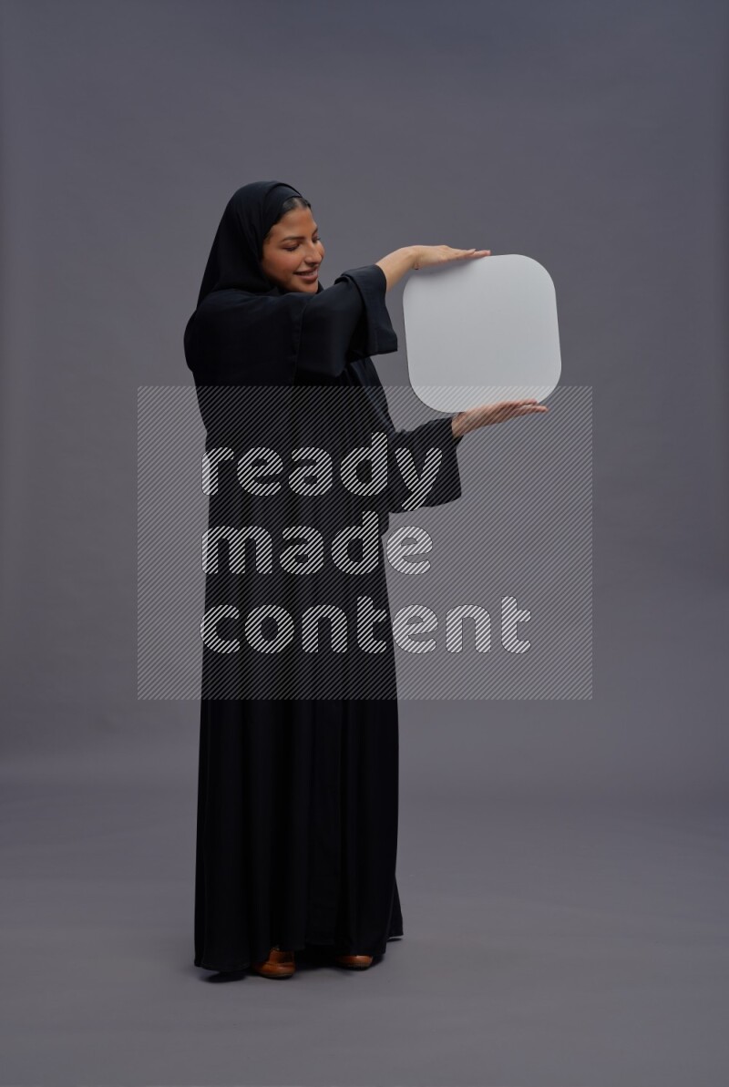 Saudi woman wearing Abaya standing holding social media sign on gray background