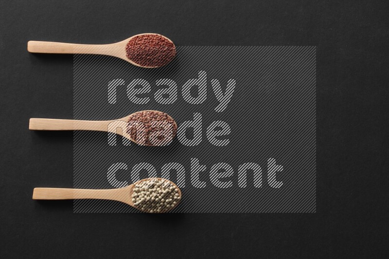 wooden spoons filled with white peppers, mustard seeds, black seeds, garden cress, cumin and flax on black flooring