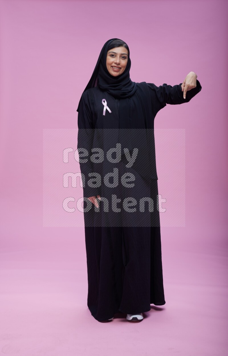 Saudi woman wearing pink ribbon on Abaya standing interacting with the camera on pink background