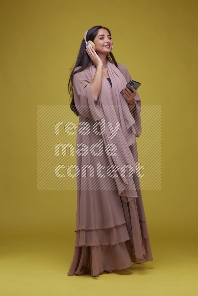 A Saudi woman Listing to Music on a Yellow Background wearing Brown Abaya