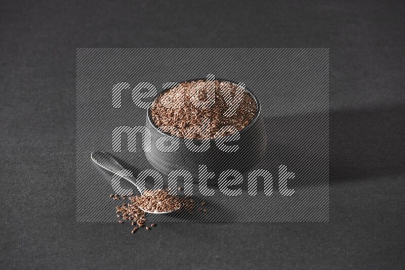 وعاء فخاري أسود ممتلئ بحبوب بذر الكتان مع ملعقة معدنية ممتلئة على أرضية سوداء