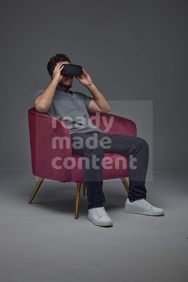 A man wearing casual and wearing VR while sitting on a burgundy chair eye level on a gray background