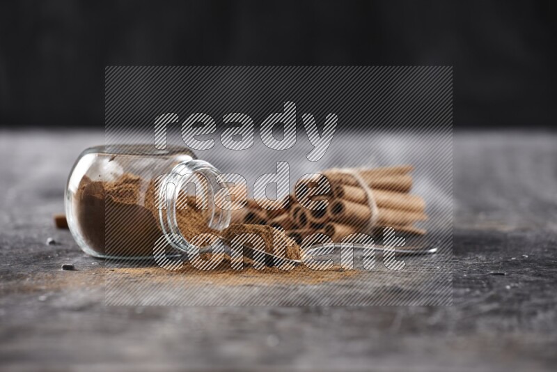 Herbal glass jar full cinnamon powder flipped and a metal spoon full of powder, cinnamon sticks stacked and bounded in the back on textured black background in different angles