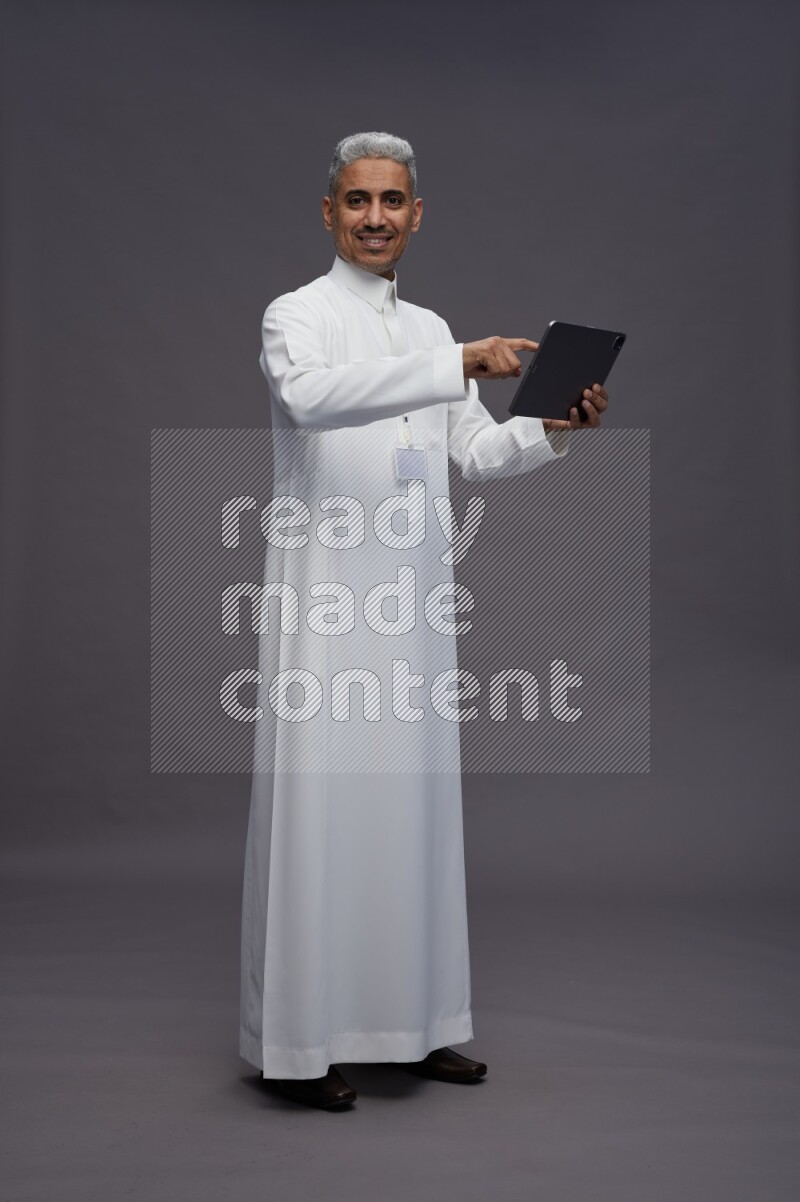 Saudi man wearing thob with neck strap employee badge standing working on tablet on gray background