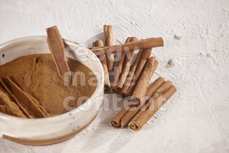 Ceramic bowl full of cinnamon powder with cinnamon sticks on the side on white background