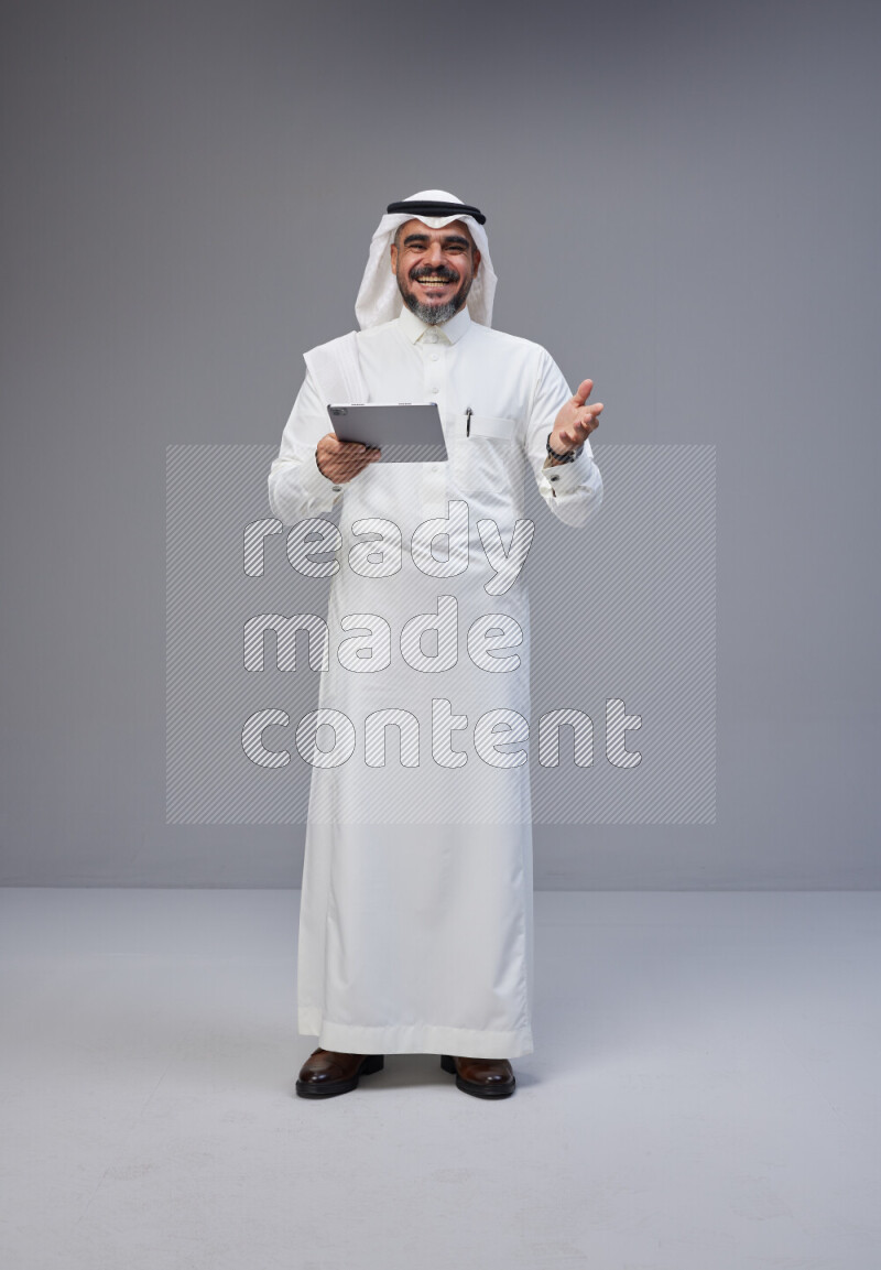 Saudi man Wearing Thob and white Shomag standing working on tablet on Gray background