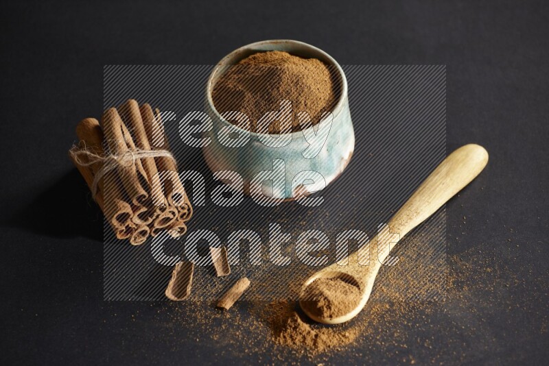 Ceramic bowl full of cinnamon powder and a wooden spoon full of powder with cinnamon sticks stacked and bounded on black background