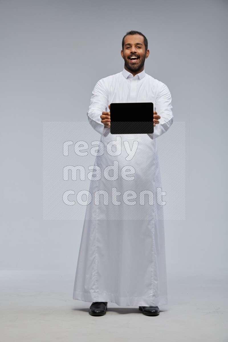 Saudi man wearing Thob standing showing tablet to camera on Gray background