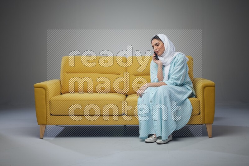 A Saudi woman wearing a light blue Abaya and a white head scarf sitting on a yellow sofa and talking in the phone while making different poses eye level on a grey background