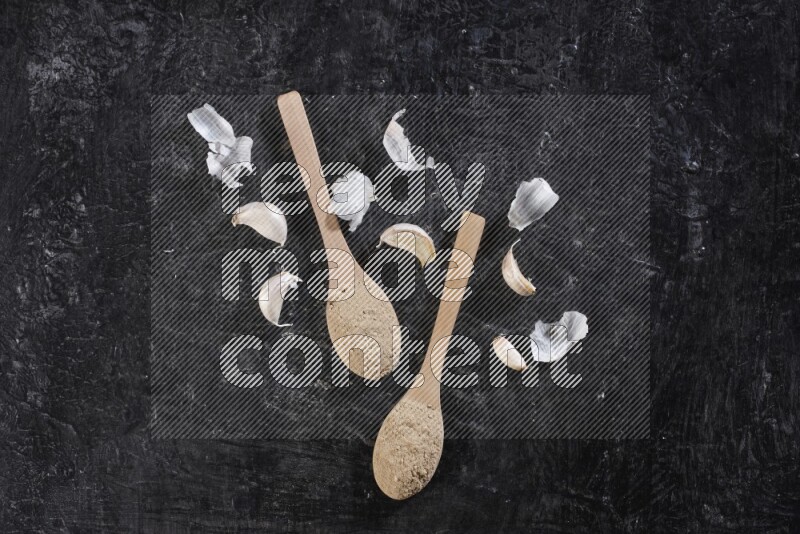 Two wooden spoons filled with garlic powder, surrounded by peeled garlic cloves and their skins on a textured black flooring