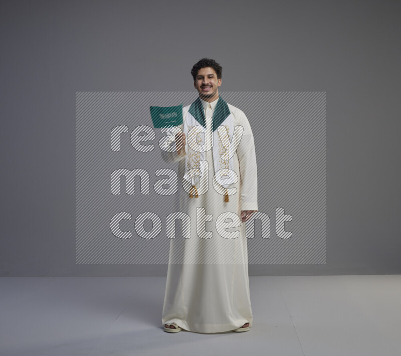 A Saudi man standing wearing thob and saudi flag scarf and holding small saudi flag on gray background