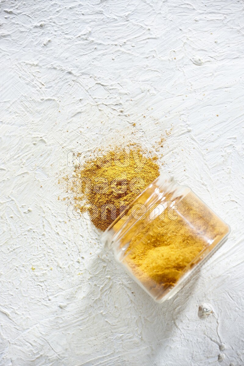 A flipped glass spice jar full of turmeric powder and powder fell out it on textured white flooring