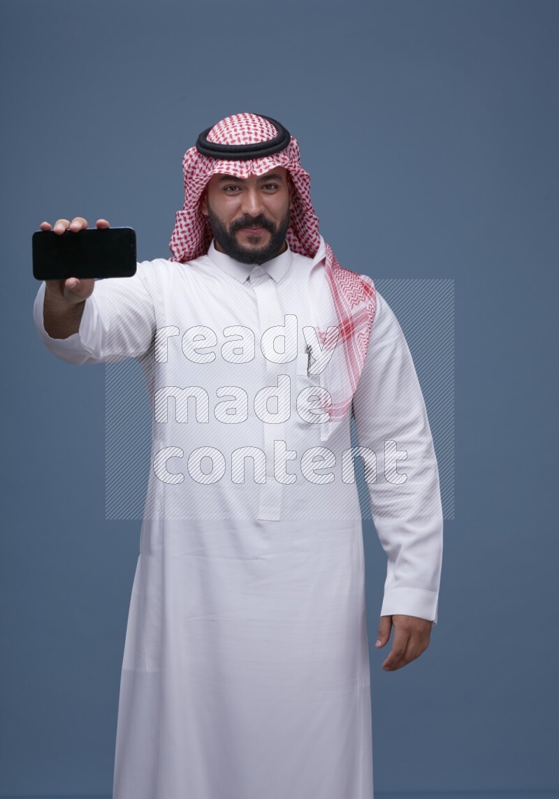 A man Showing his smartphone Screen on Blue Background wearing Saudi Thob