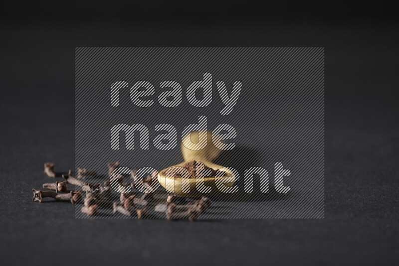 A wooden spoon full of cloves powder with spreaded whole cloves on a black flooring