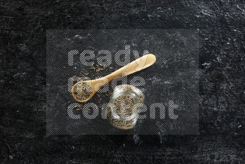 A glass spice jar and a wooden spoon full of cumin seeds on a textured black flooring