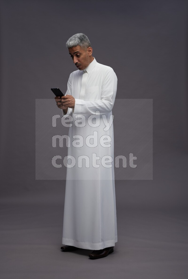 Saudi man wearing thob standing texting on phone on gray background