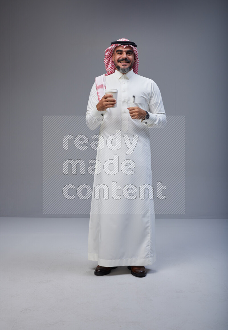 Saudi man Wearing Thob and red Shomag standing holding paper cup on Gray background