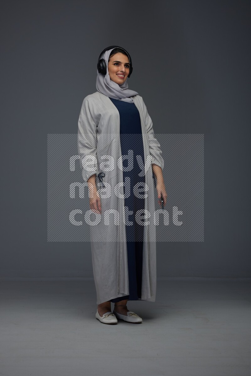 A Saudi woman wearing a light gray Abaya and head scarf standing and listening to music on a grey background