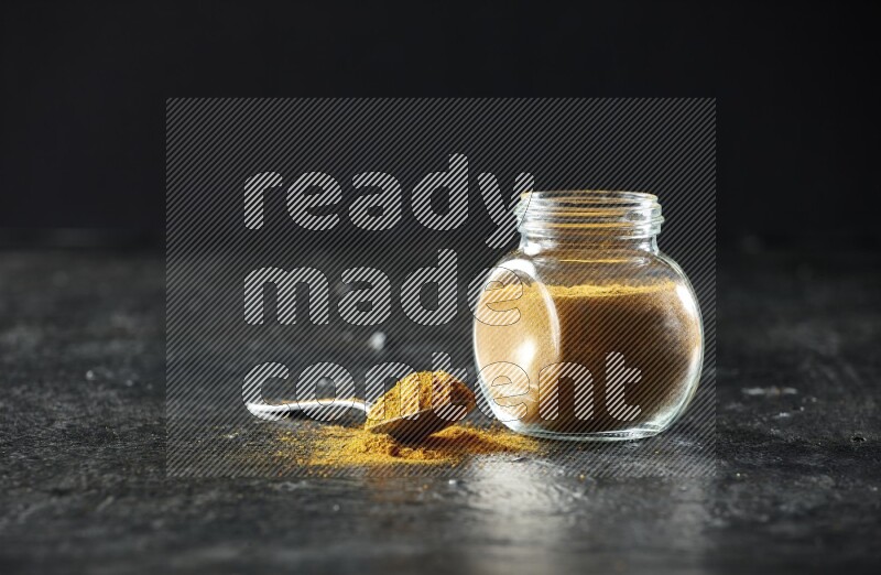 A glass spice jar and metal spoon full of turmeric powder on textured black flooring