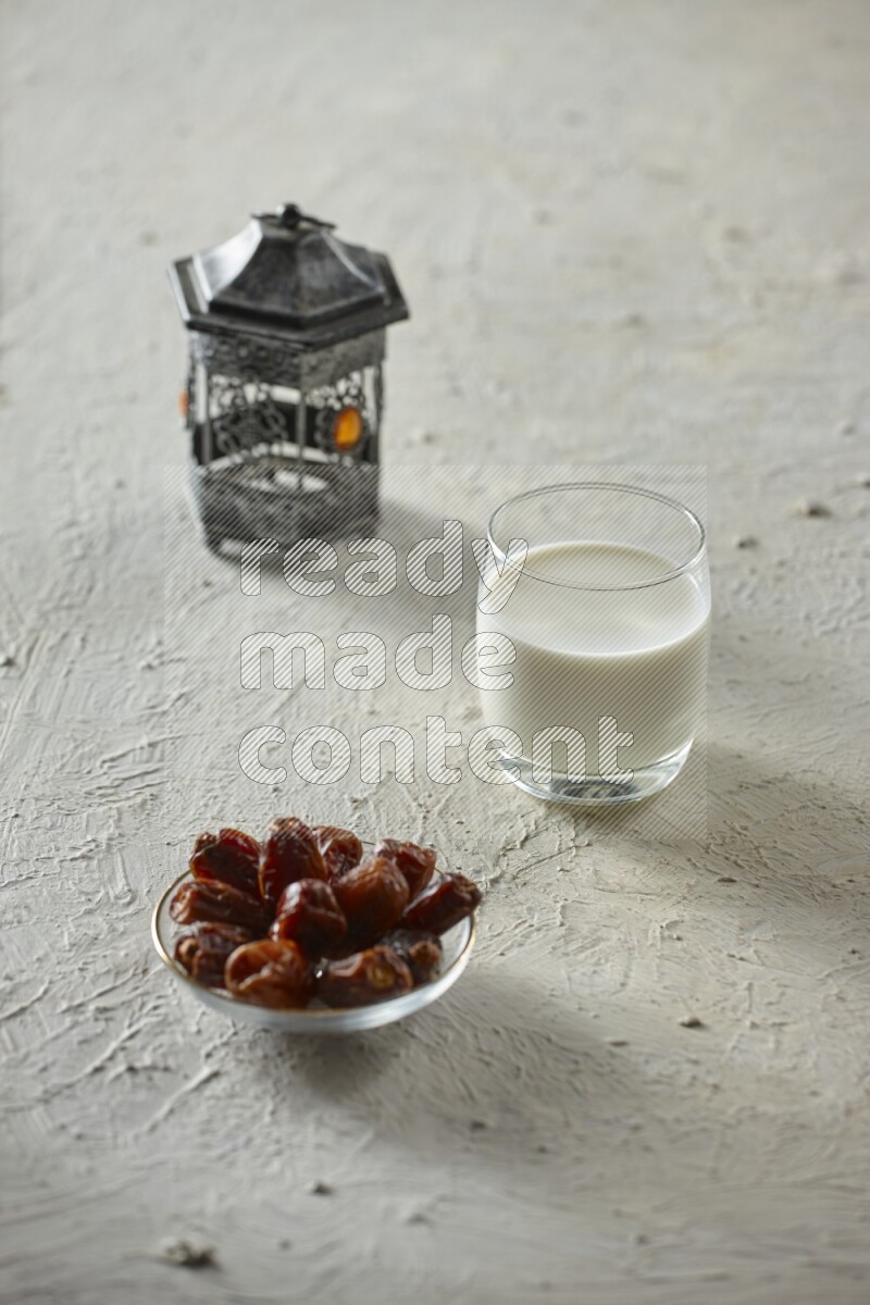 A silver lantern with different drinks, dates, nuts, prayer beads and quran on textured white background
