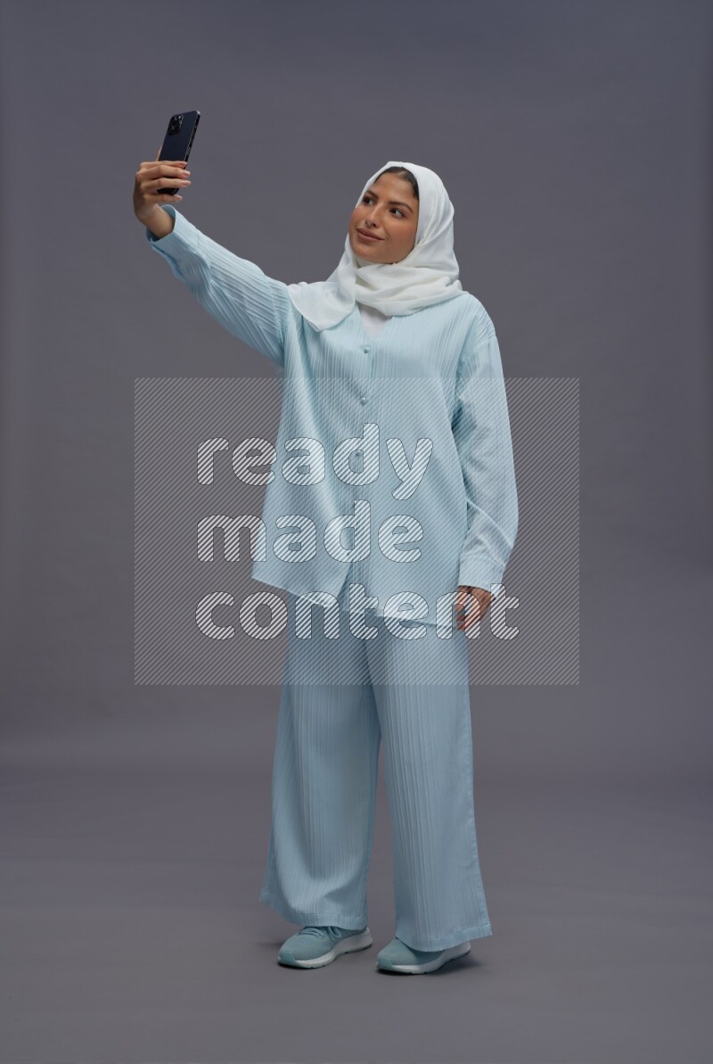 Saudi woman wearing hijab clothes standing taking selfie on gray background