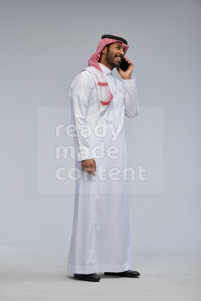 Saudi man Wearing Thob and shomag standing talking on phone on Gray background