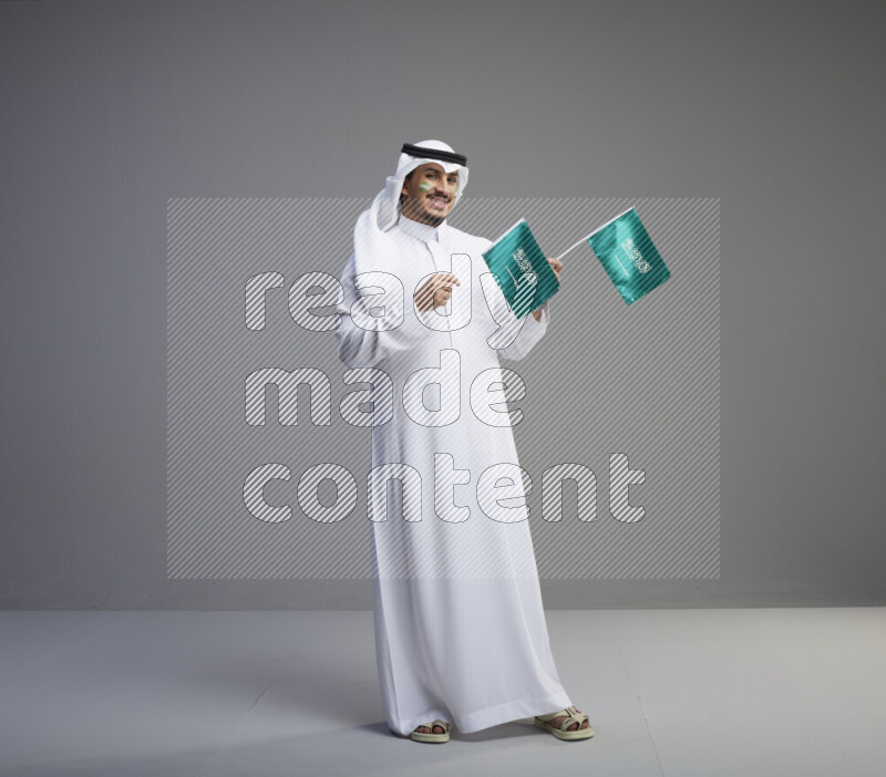 A Saudi man standing wearing thob and white shomag with face painting raising small saudi flag on gray background