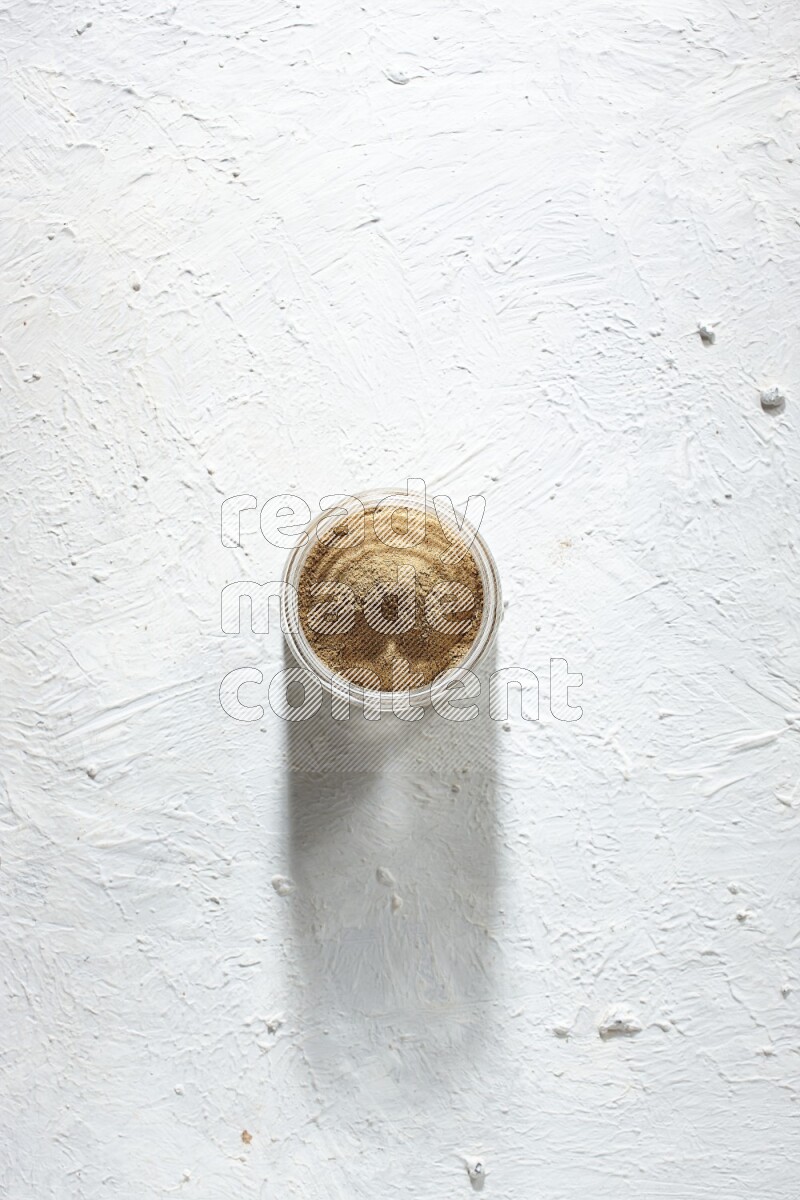 A glass jar full of cumin powder on textured white flooring