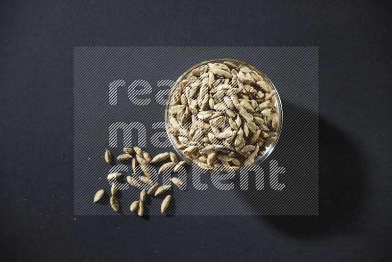 A glass bowl full of cardamom and more seeds spreaded beneath the bowl on black flooring