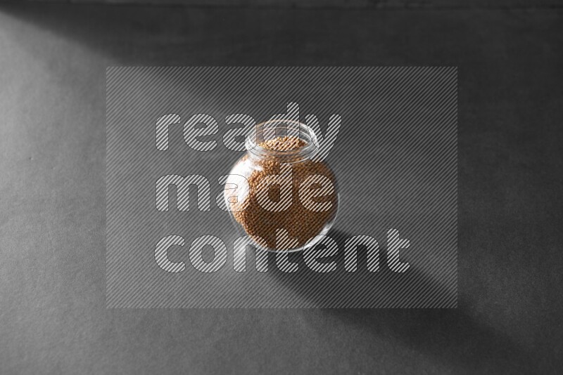 A glass spice jar full of mustard seeds on black flooring